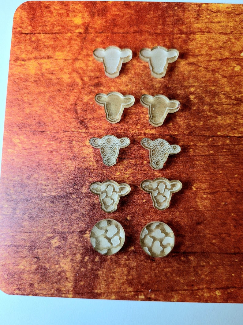 Peut inclure: Un ensemble de huit boucles d'oreilles en bois en forme de vache. Les boucles d'oreilles sont de styles vari&eacute;s, notamment en blanc uni, en brun et avec un motif. Les boucles d'oreilles sont pr&eacute;sent&eacute;es sur une surface en bois.