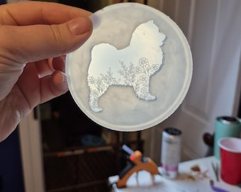 May include: A clear, round coaster featuring a detailed silhouette of a dog breed, adorned with floral accents. The dog's outline is filled with delicate flower and leaf patterns, creating a decorative and artistic design. The coaster is held up by a hand.