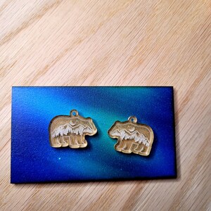May include: Two bear-shaped charms made of clear acrylic, each featuring an etched white design of a mountain range, pine forest, and a crescent moon. The charms are positioned on a rectangular card with a blue and green gradient background.