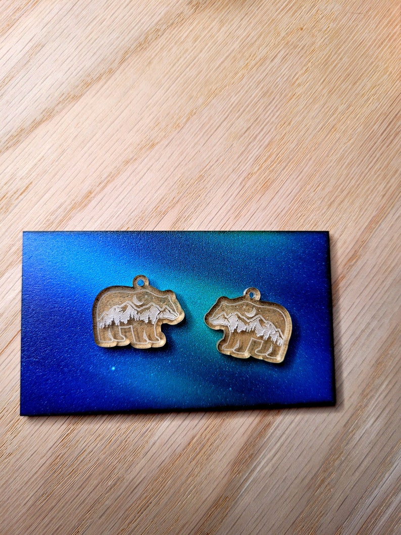 May include: Two bear-shaped charms made of clear acrylic, each featuring an etched white design of a mountain range, pine forest, and a crescent moon. The charms are positioned on a rectangular card with a blue and green gradient background.