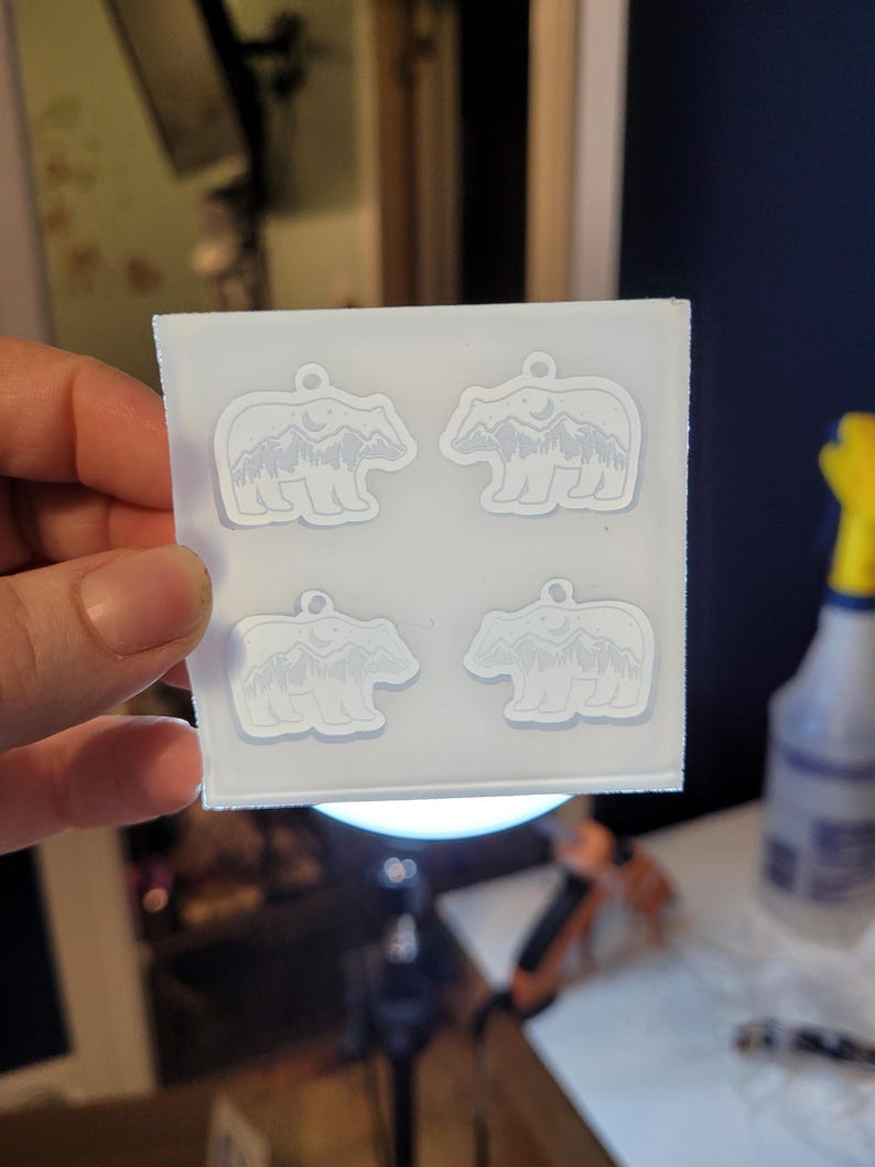 May include: A square, translucent silicone mould featuring four identical cavities shaped like bears. Each bear cavity contains an embossed design of a mountain range, a forest of pine trees, and a crescent moon within the bear's silhouette.