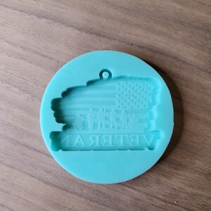 May include: A teal blue silicone mould for resin crafts. The mould features an American flag with a silhouette of soldiers in front of it. The text "Veteran" is written below the flag.