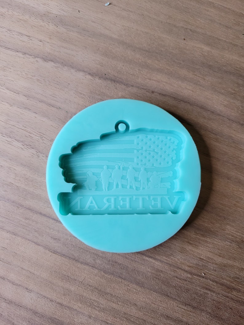 May include: A teal blue silicone mould for resin crafts. The mould features an American flag with a silhouette of soldiers in front of it. The text "Veteran" is written below the flag.
