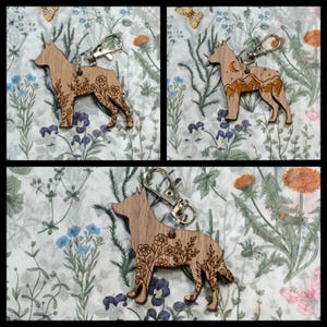 May include: Three wooden dog-shaped keychains with silver-colored clasps. One keychain features floral designs, another has a mountain and moon design, and the third has floral designs. The keychains are displayed on a floral patterned background.