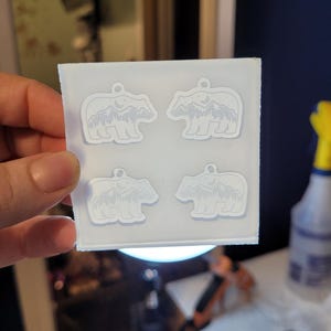 May include: A square, translucent silicone mould featuring four identical cavities shaped like bears. Each bear cavity contains an embossed design of a mountain range, a forest of pine trees, and a crescent moon within the bear's silhouette.