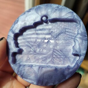 May include: A round, iridescent purple and white bauble with a raised image of a flag and soldiers. The bauble has a hole at the top for hanging.