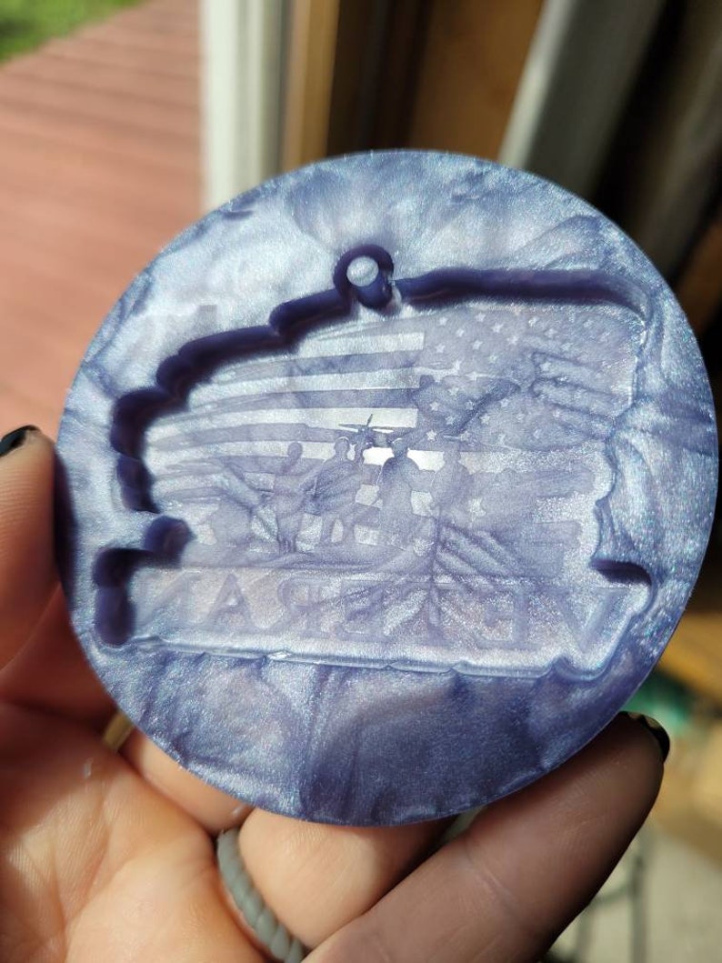 May include: A round, iridescent purple and white bauble with a raised image of a flag and soldiers. The bauble has a hole at the top for hanging.