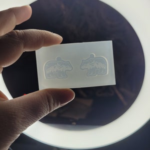 May include: A translucent silicone mould featuring two identical bear-shaped cavities. Each bear silhouette contains an intricate recessed design of a mountain range, a forest of pine trees, and a crescent moon within the upper body area.
