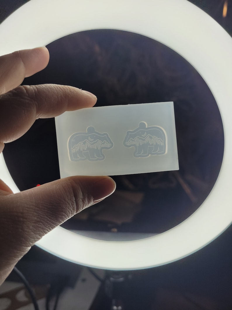 May include: A translucent silicone mould featuring two identical bear-shaped cavities. Each bear silhouette contains an intricate recessed design of a mountain range, a forest of pine trees, and a crescent moon within the upper body area.