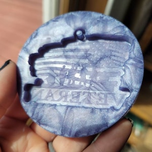 May include: A round, purple and white, iridescent, resin ornament with a silhouette of a flag and the word "Veteran" in the centre.