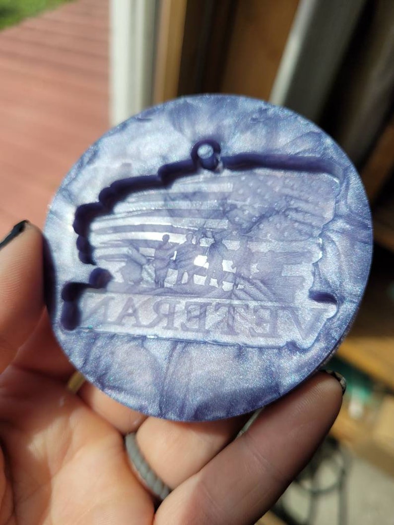May include: A round, purple and white, iridescent, resin ornament with a silhouette of a flag and the word "Veteran" in the centre.