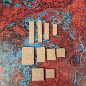 May include: A collection of small, rectangular, and square wooden blocks with measurements in inches. The blocks are various sizes, with dimensions such as 2 x .5 inches, 1.5 x 1 inches, and 1 x 1 inch. The blocks are arranged on a textured, multi-colored surface.