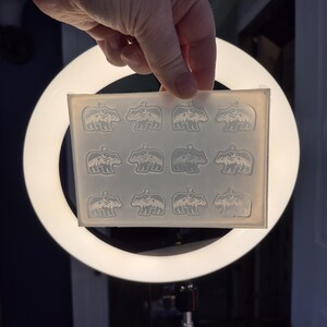 May include: A translucent silicone mould featuring twelve identical cavities shaped like a bear silhouette with a mountain range and moon design inside. The rectangular mould is flexible and clear, designed for casting small decorative pendants.