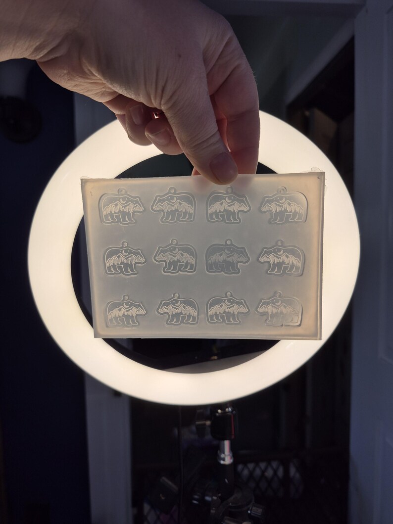 May include: A translucent silicone mould featuring twelve identical cavities shaped like a bear silhouette with a mountain range and moon design inside. The rectangular mould is flexible and clear, designed for casting small decorative pendants.
