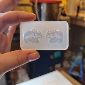 May include: A rectangular, translucent silicone mould featuring two identical bear-shaped cavities. Each bear cavity contains an embossed design of a mountain range, a crescent moon, and stars, with a small circular hole at the top for a pendant loop.