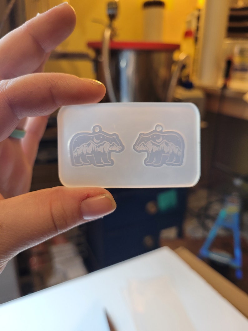 May include: A rectangular, translucent silicone mould featuring two identical bear-shaped cavities. Each bear cavity contains an embossed design of a mountain range, a crescent moon, and stars, with a small circular hole at the top for a pendant loop.