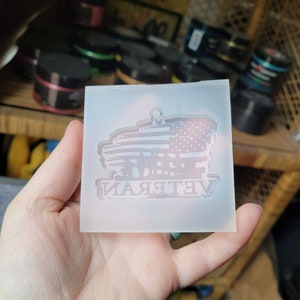 May include: A clear silicone mould for resin casting. The mould features a design of an American flag with the word "Veteran" underneath. The flag is in the shape of a banner with a silhouette of soldiers standing in front of it.