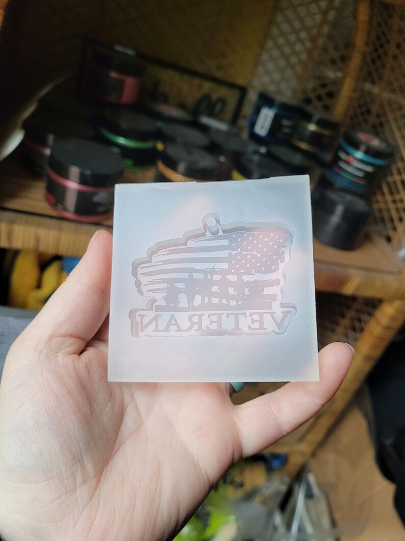 May include: A clear silicone mould for resin casting. The mould features a design of an American flag with the word "Veteran" underneath. The flag is in the shape of a banner with a silhouette of soldiers standing in front of it.