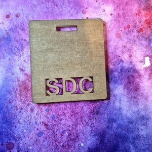 May include: A wooden tag with the letters "SDC" cut out. The tag is brown and has a hole at the top for hanging.