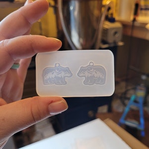 May include: A rectangular, translucent silicone mould featuring two identical bear-shaped cavities. Each bear silhouette contains an internal design of a mountain range, a crescent moon, and stars, with a small circular hole at the top for a pendant.