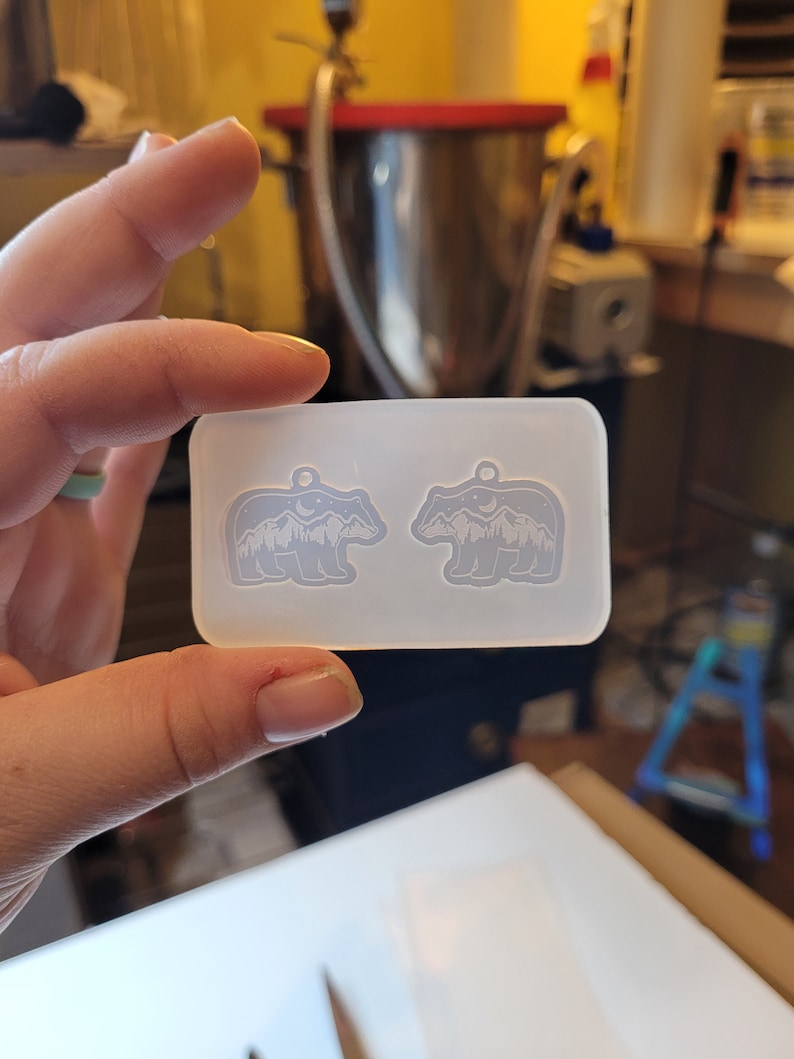 May include: A rectangular, translucent silicone mould featuring two identical bear-shaped cavities. Each bear silhouette contains an internal design of a mountain range, a crescent moon, and stars, with a small circular hole at the top for a pendant.