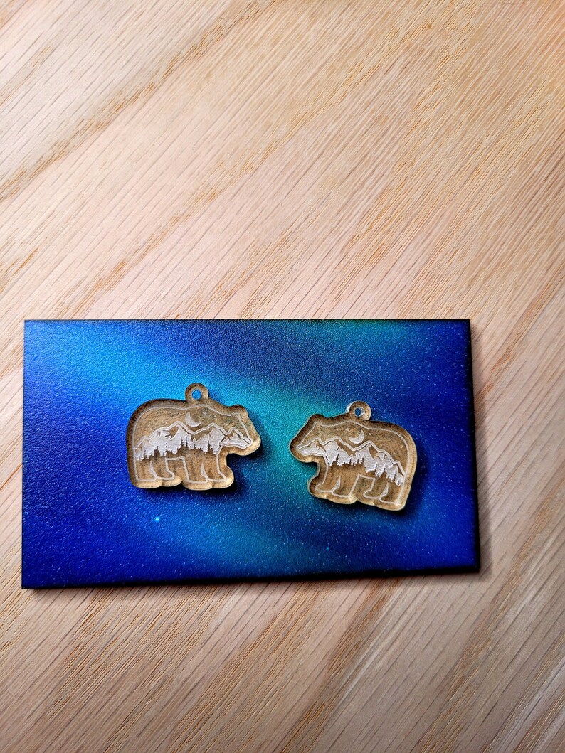 May include: Two clear acrylic bear-shaped pendants featuring an etched design of a mountain range, a forest of pine trees, and a crescent moon. Each bear charm has a small circular hole at the top for attachment to a necklace or jewellery chain.