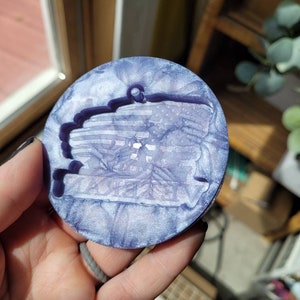 May include: A round, purple and white resin coaster with a raised, textured design. The coaster has a slightly iridescent finish.