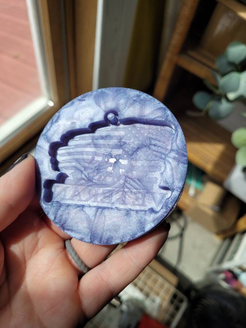 May include: A round, purple and white resin coaster with a raised, textured design. The coaster has a slightly iridescent finish.