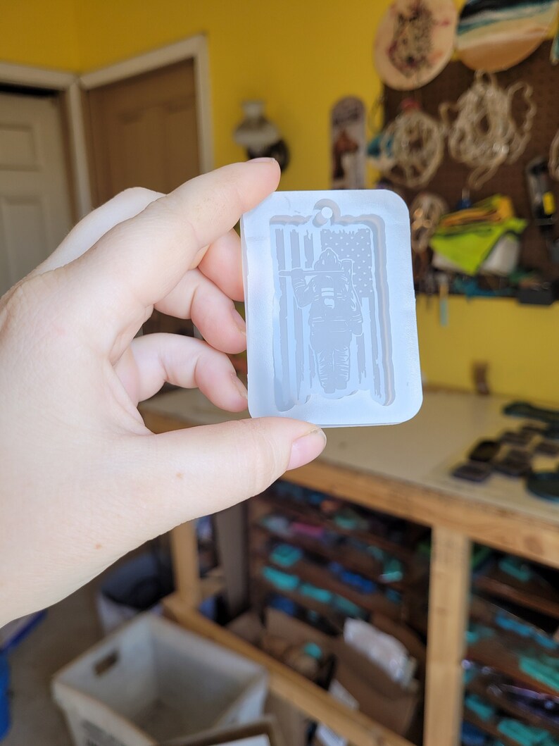 May include: A clear silicone mould for resin casting, featuring a firefighter standing in front of an American flag. The mould is designed to create a keychain or pendant.