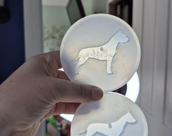 May include: Two round, translucent coasters with a dog silhouette design. The dog is filled with a floral pattern. The coasters are held in front of a bright light, and a blue cup is visible in the background.