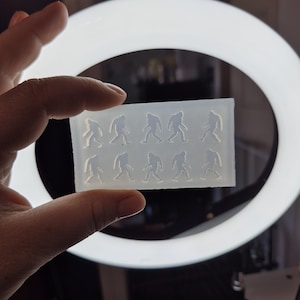 May include: A clear silicone mold with 10 impressions of a Bigfoot silhouette. The mold is held in a hand against a white ring light.