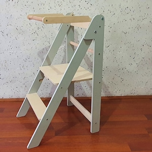 May include: A light green and natural wood step stool with a small table. The stool has a ladder-like design with a seat and a small table at the top. The wood is light-coloured.