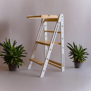 May include: White wooden step stool with a safety rail and two shelves. The stool is made of wood and has a white finish. The stool is standing on a light gray surface.