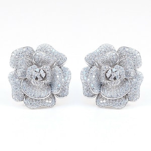 May include: A pair of silver-toned flower-shaped stud earrings. Each earring is designed as a rose, with multiple layers of petals. The petals and center are encrusted with small, sparkling stones, creating a shimmering effect. The earrings are set against a white background.
