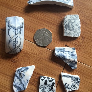 May include: A collection of seven blue and white porcelain shards with floral patterns. The shards are arranged on a wooden surface with a British one pound coin for scale.