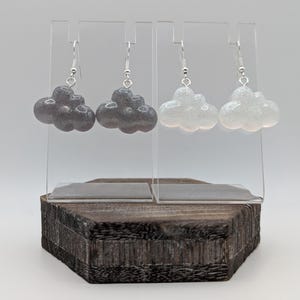 May include: Two pairs of cloud-shaped earrings on a wooden display stand. One pair is dark grey, the other white, both with silver hooks. The earrings have a glittery texture, resembling fluffy clouds. A charming accessory.