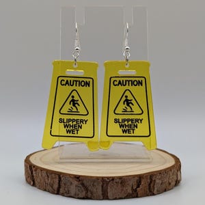 May include: A pair of yellow caution sign earrings with the text "CAUTION SLIPPERY WHEN WET" and a black silhouette of a person slipping.
