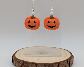 May include: A pair of Halloween pumpkin earrings. Each earring has a small, orange pumpkin with a carved face and a green stem. The earrings hang from silver-coloured hooks. The pumpkins are displayed on a clear stand.