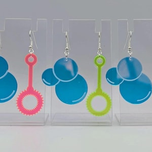May include: A set of earrings with a bubble design. Each earring has three blue bubble shapes and a colourful bubble wand. The wands are pink, green, and yellow. The earrings hang from silver hooks.