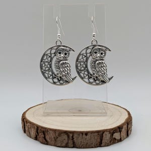 May include: Silver owl earrings. Each earring features an owl perched on a crescent moon with a decorative filigree design. The earrings are displayed on a clear stand atop a wooden slice.