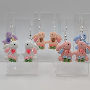 May include: A selection of bunny-shaped earrings in pastel shades. The earrings feature a bow and floral details, with colours including purple, pink, blue, and peach. They are displayed on clear stands, showcasing the Easter-themed jewellery.