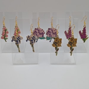 May include: A selection of floral earrings with gold-coloured hooks. The earrings showcase diverse flower designs, including pink, purple, and yellow blooms. Each earring is displayed on a clear acrylic stand, highlighting the intricate details of the jewellery.