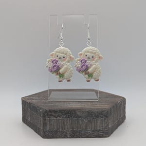 May include: A pair of earrings depicting white sheep holding purple flower bouquets. The sheep have pink cheeks and are suspended from silver hooks. The earrings are displayed on a clear acrylic stand.