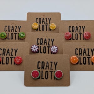 May include: A selection of fruit-shaped stud earrings presented on brown card tags. The earrings depict kiwi, orange, watermelon, and other fruits. The tags are printed with the text "CRAZY LOTIO".