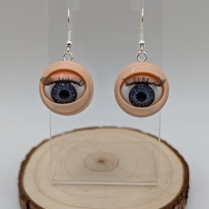 May include: A pair of dangle earrings featuring blue eyes with black eyelashes and a flesh-toned surrounding. The earrings are made of polymer clay and have silver hooks.