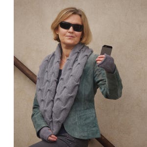 May include: A woman wearing a gray cable knit scarf and a green suede jacket. She is wearing sunglasses and holding a black smartphone.