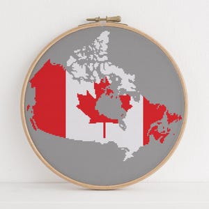 May include: A round embroidery hoop featuring a gray background with a map of Canada in white and red. The Canadian flag is prominently displayed. The wooden hoop is set against a white background.