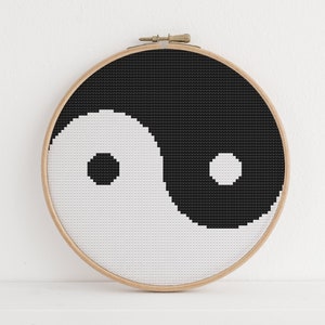 May include: A round, cross-stitch embroidery hoop featuring a black and white Yin and Yang symbol. The design is centered within a wooden hoop, creating a balanced and minimalist aesthetic. The background is a plain white surface.