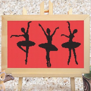 May include: A framed cross-stitch artwork showing three black ballerina silhouettes against a red backdrop. The artwork is displayed on a small wooden easel, with potted plants on either side.
