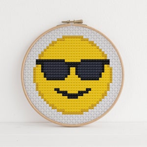 May include: A yellow smiley face with black sunglasses embroidered on a white canvas in a wooden embroidery hoop.